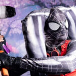 A cosplayer in a black and red Spider-Man inspired suit swinging a hammer prop at rAge
