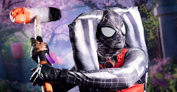 A cosplayer in a black and red Spider-Man inspired suit swinging a hammer prop at rAge